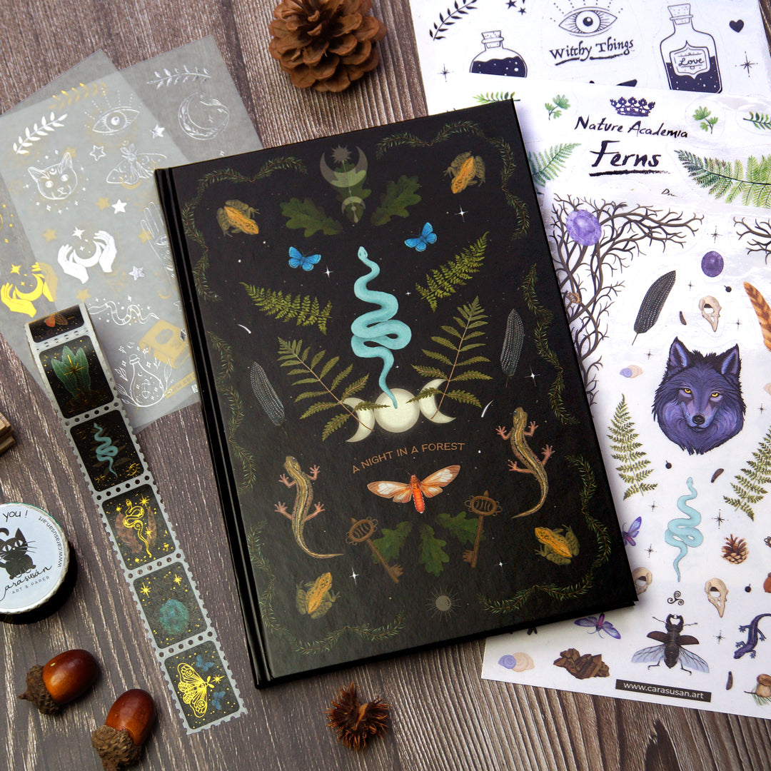 A witchy journaling set displayed on a wooden surface, featuring a dark forest-themed journal titled "A Night in a Forest" with intricate illustrations of a snake, ferns, butterflies, lizards, and other forest elements. The set includes matching stickers with various designs, such as leaves, animals, and mystical symbols, and a roll of washi tape with similar forest and mystical motifs. 