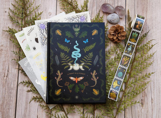 A witchy journaling set displayed on a wooden surface, featuring a dark forest-themed journal titled "A Night in a Forest" with intricate illustrations of a snake, ferns, butterflies, lizards, and other forest elements. The set includes matching stickers with various designs, such as leaves, animals, and mystical symbols, and a roll of washi tape with similar forest and mystical motifs. The scene is decorated with pinecones, stones, and sprigs of greenery, enhancing the natural and magical theme.